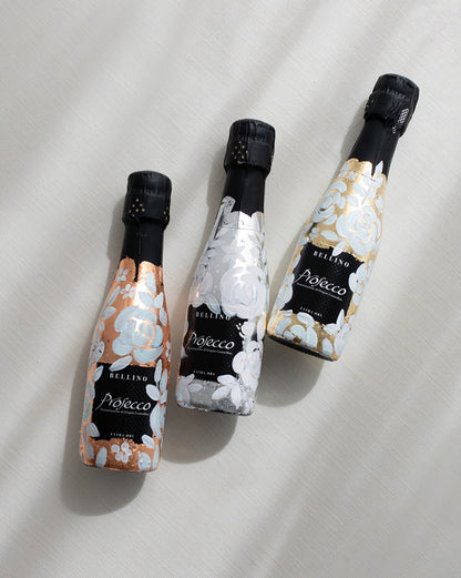 Mini bottles of Prosecco hand painted with a white roses design on a metallic base. Gold, silver and copper examples of the same design. The perfect luxury bridesmaid proposal gift. Fancy Folk Design
