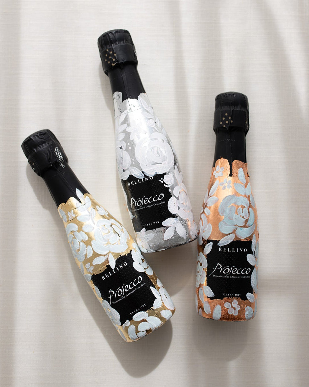 Mini bottles of Prosecco hand painted with a white roses design on a metallic base. Gold, silver and copper examples of the same design. The perfect luxury bridesmaid proposal gift. Fancy Folk Design