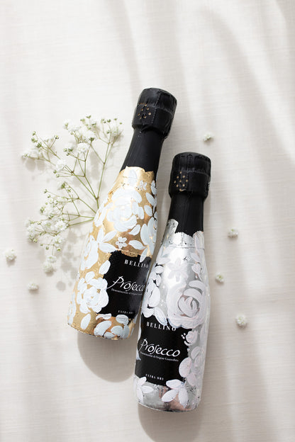 Mini bottles of Prosecco hand painted with a white roses design on a metallic gold and silver leaf base. The perfect luxury bridesmaid proposal gift. Fancy Folk Design