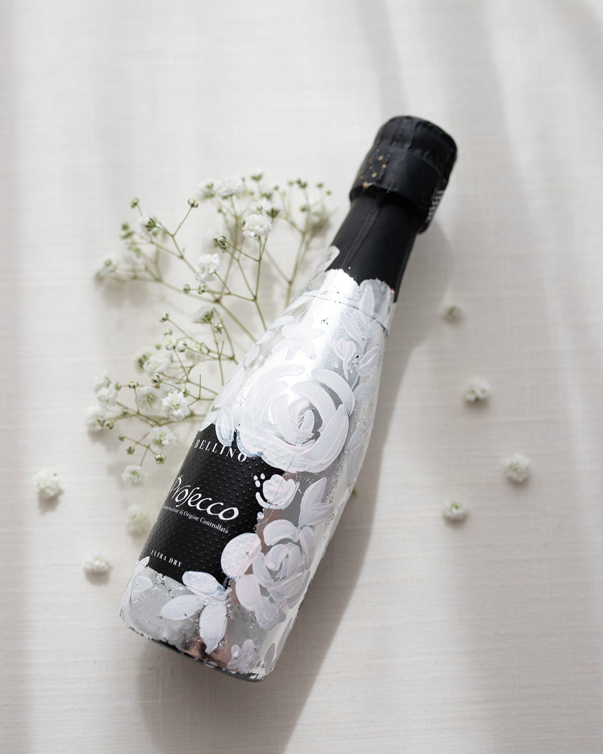 Mini bottles of Prosecco hand painted with a white roses design on a metallic silver leaf base. The perfect luxury bridesmaid proposal gift. Fancy Folk Design