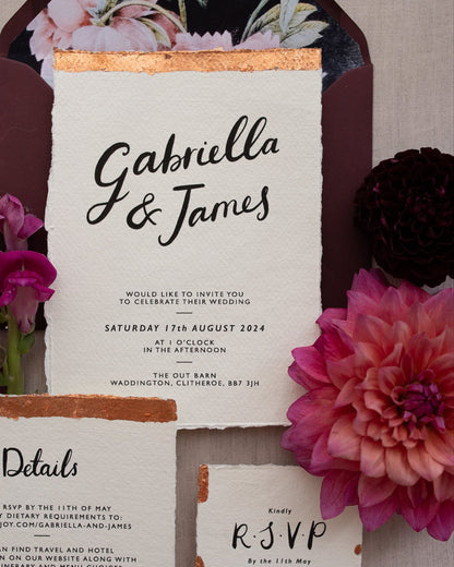 Close up of a Modern Calligraphy Wedding Invitation, Details Card and RSVP with rich burgundy coloured envelopes. Our wedding stationery features a beautiful hand lettered calligraphy names design printed on textured handmade Indian paper with hand torn edges for a soft romantic feel. Each piece is lovingly hand finished with copper leaf for a luxury feel. Styled with burgundy and coral florals, perfect for autumn and winter weddings. Fancy Folk