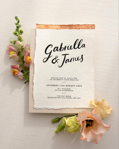 Modern Calligraphy Wedding Invitation, with neutral almond coloured envelope. Features a beautiful hand lettered calligraphy names design printed on textured handmade Indian paper with hand torn edges for a soft romantic feel. Each piece is lovingly hand finished with copper leaf for a luxury feel. Styled with peachy florals. Fancy Folk