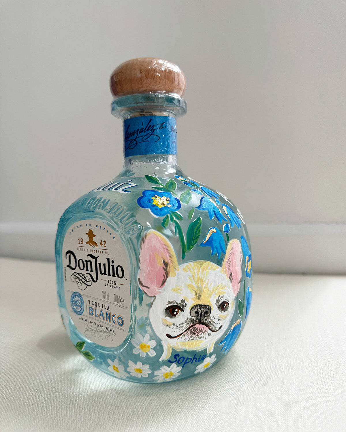 Hand painted pet portrait of a cute french bulldog on a celebration bottle of   alcohol. A pale blue bottle is decorated with beautiful blue florals and calligraphy pets name. Finished with accents of gold leaf. Perfect personalised gift. Fancy Folk
