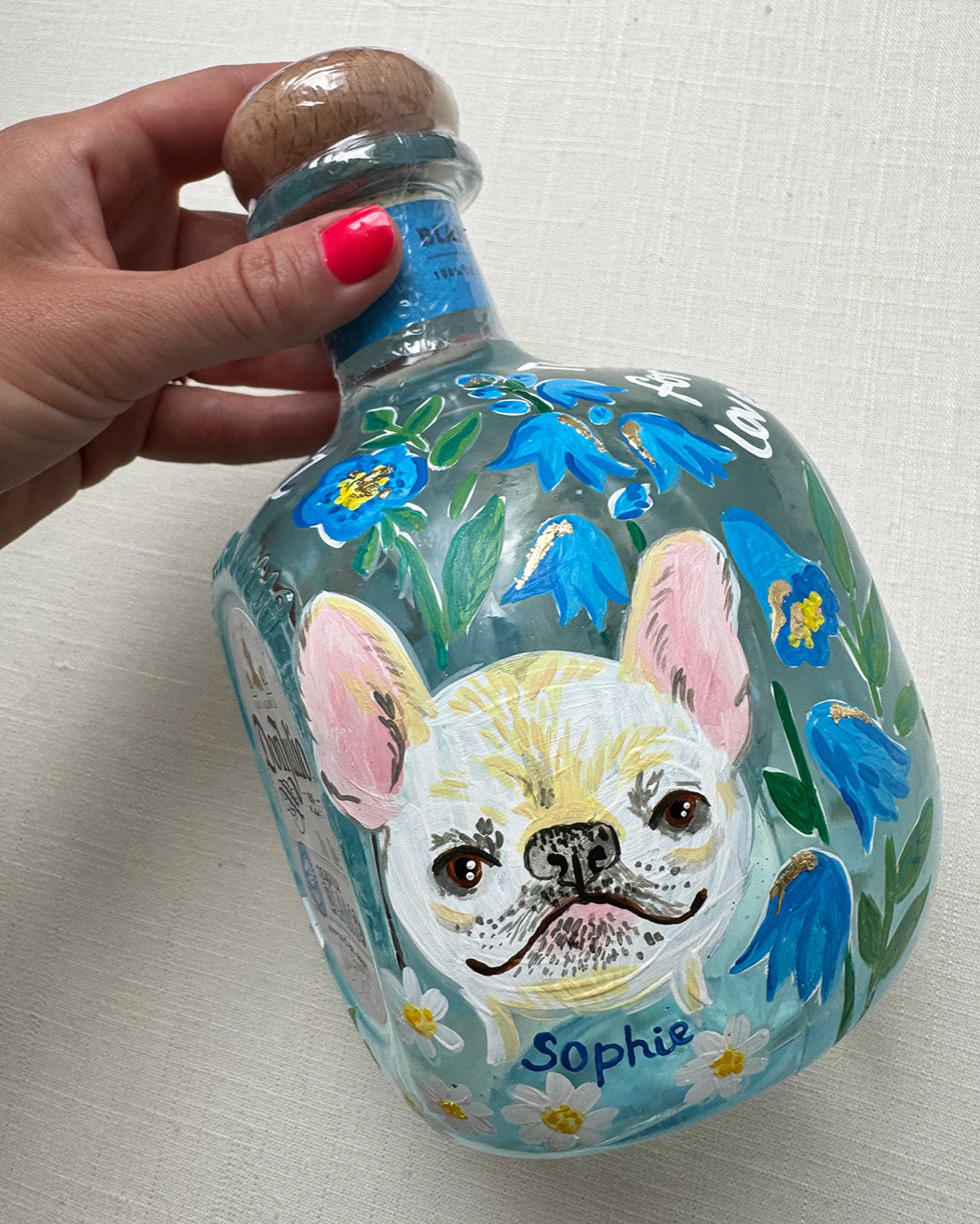 Hand painted pet portrait of a cute french bulldog on a celebration bottle of   alcohol. A pale blue bottle is decorated with beautiful blue florals and calligraphy pets name. Finished with accents of gold leaf. Perfect personalised gift. Fancy Folk