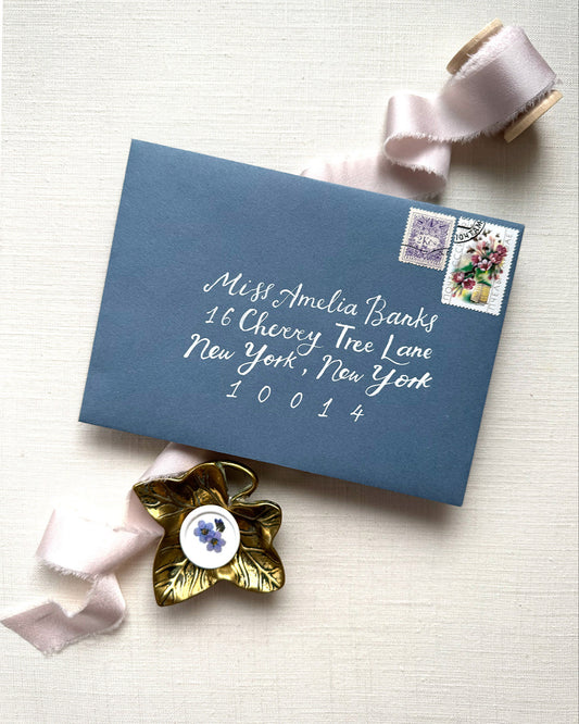 Wedding envelope calligraphy names and address in a white ink. Written on a dusty blue envelope. Fancy Folk