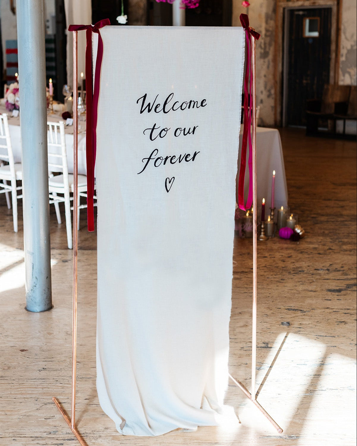 Fabric wedding welcome sign, hand painted quote in a modern calligraphy style onto a neutral cotton linen fabric.
Hanging from a copper frame with velvet ribbon bows either side to greet your wedding guests. Fancy Folk