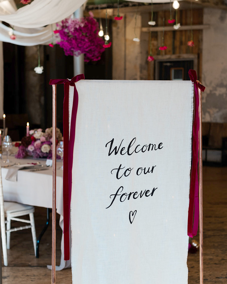 Fabric wedding welcome sign, hand painted quote in a modern calligraphy style onto a neutral cotton linen fabric.
Hanging from a copper frame with velvet ribbon bows either side to greet your wedding guests. Fancy Folk
