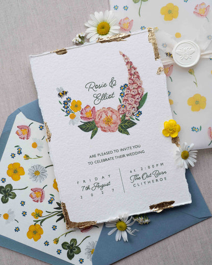 Watercolour hand painted florals of meadow flowers and a little bee make up a posy. Couples names are nestled in the floral. Printed on handmade Indian cotton paper with gold leaf accent. Styled with white bee wax seals, floral envelope liners and silk ribbon. Dusty blue envelopes and floral vellum wrap. 