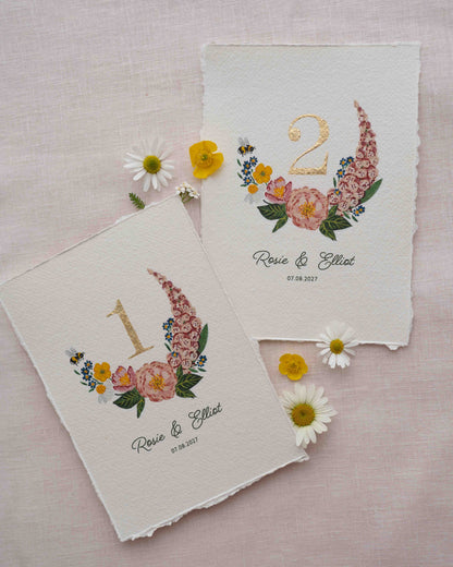 Pretty wild flower wedding table number. With gold leaf number in the centre of the posy. Little bee flying into the flowers. Couples names and wedding date printed below. Styled with fresh floewers on linen.