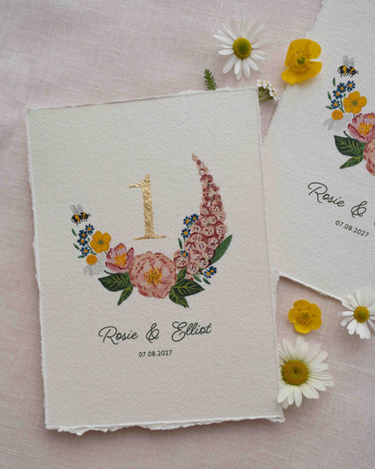 Pretty wild flower wedding table number. With gold leaf number in the centre of the posy. Little bee flying into the flowers. Couples names and wedding date printed below. Styled with fresh floewers on linen.