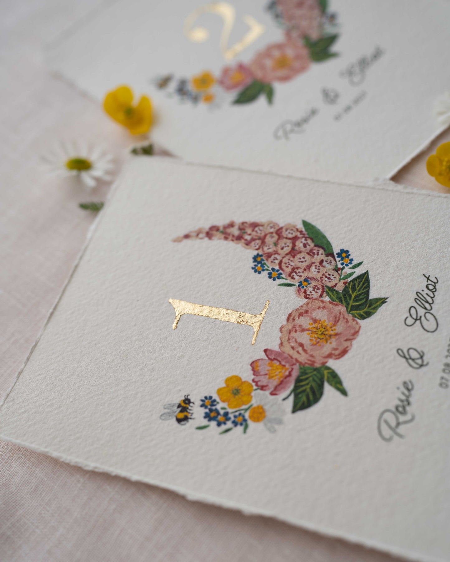 Pretty wild flower wedding table number. With gold leaf number in the centre of the posy. Little bee flying into the flowers. Couples names and wedding date printed below. Styled with fresh floewers on linen.