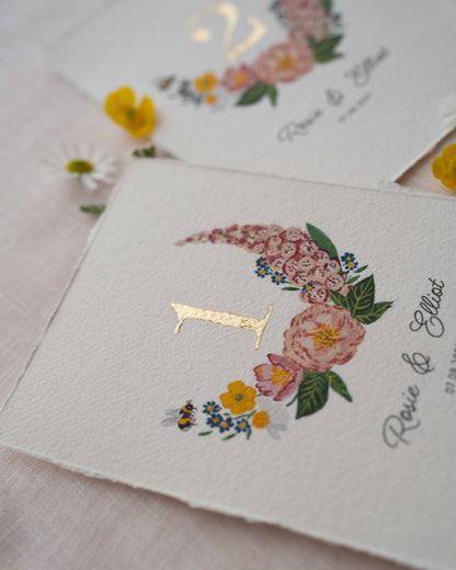 Pretty wild flower wedding table number. With gold leaf number in the centre of the posy. Little bee flying into the flowers. Couples names and wedding date printed below. Styled with fresh floewers on linen.