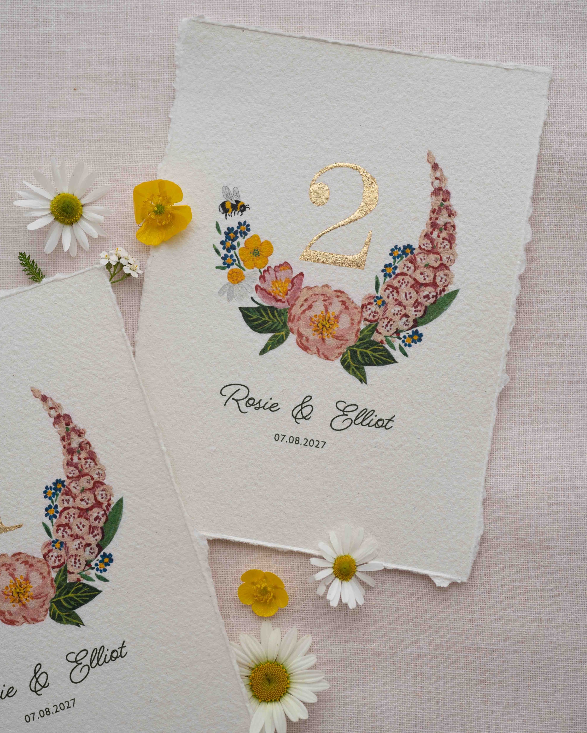 number. With gold leaf number in the centre of the posy. Little bee flying into the flowers. Couples names and wedding date printed below. Styled with fresh floewers on linen.