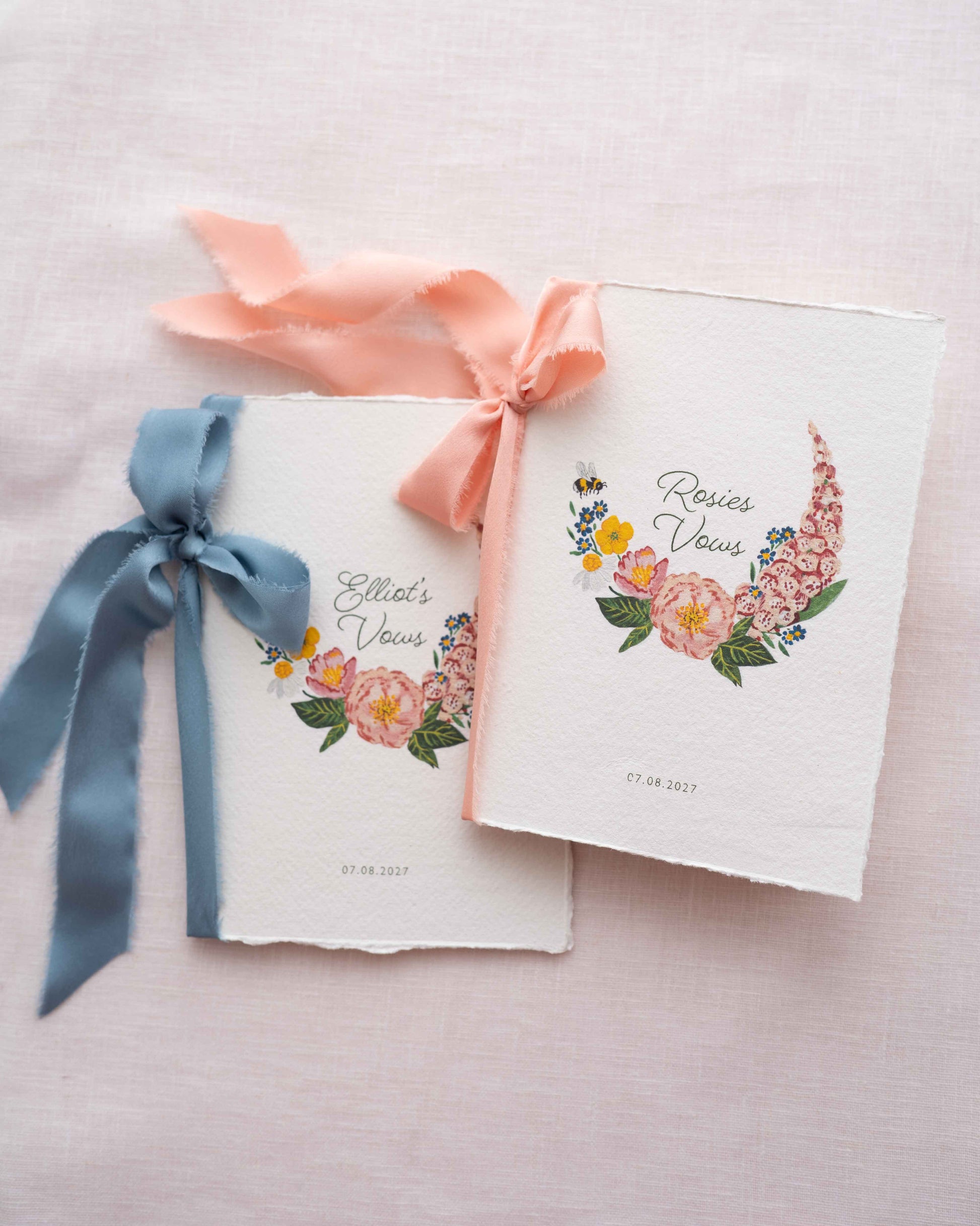 Two floral couples vow book with silk ribbons on a light pink background. Watercolour hand painted florals of meadow flowers and a little bee make up a posy. Couples names are nestled in the floral and printed wedding date below. Printed on handmade Indian cotton paper, tied silk ribbon bow. Fancy Folk Design