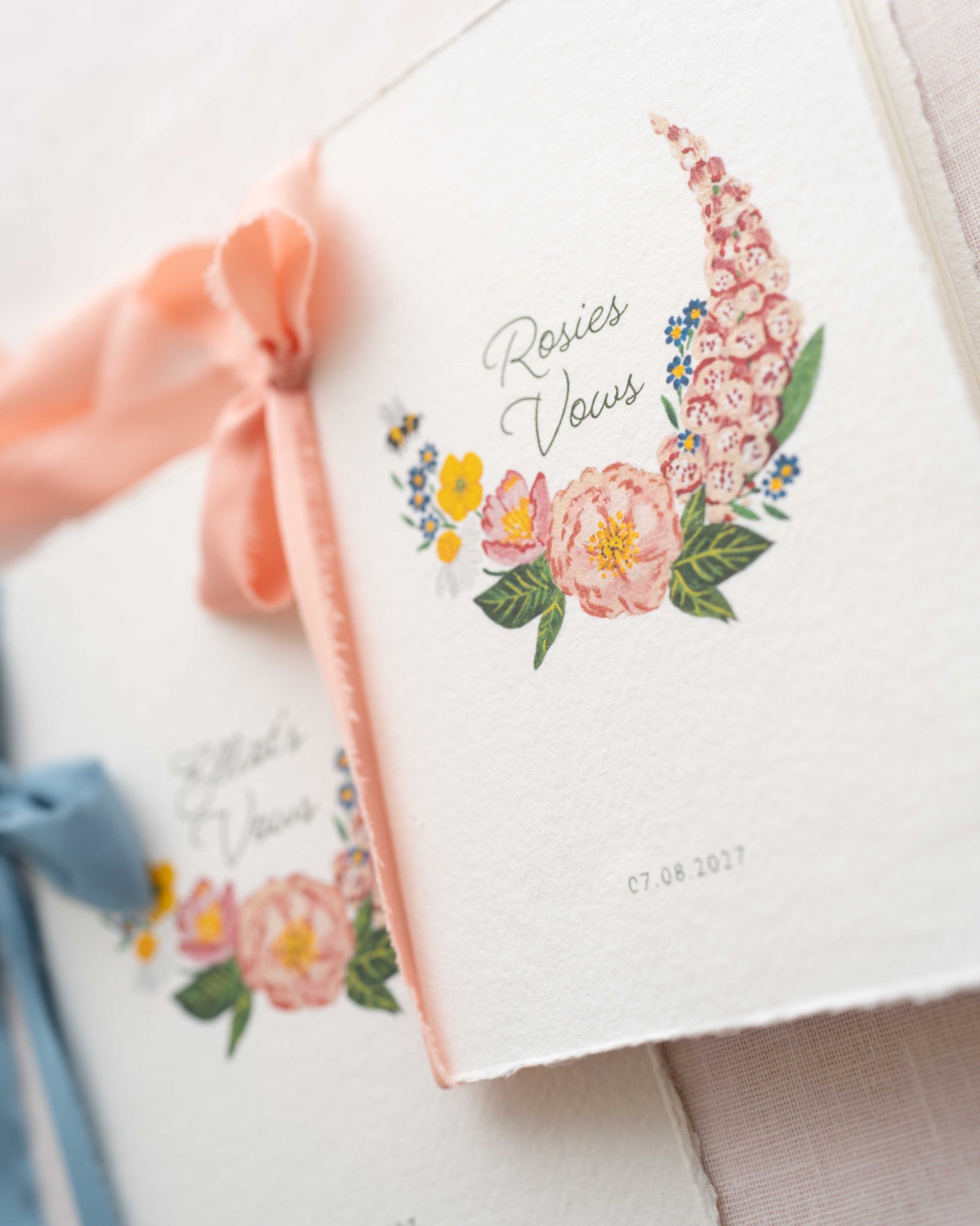 Close up of Wild Flower Wedding Vow Book which reads Rosies Vows. The text is nestled in a watercolour posy with a little bumble bee flying into the design. Printed on handmade Indian cotton paper with blush pink ribbon. Fancy Folk Design