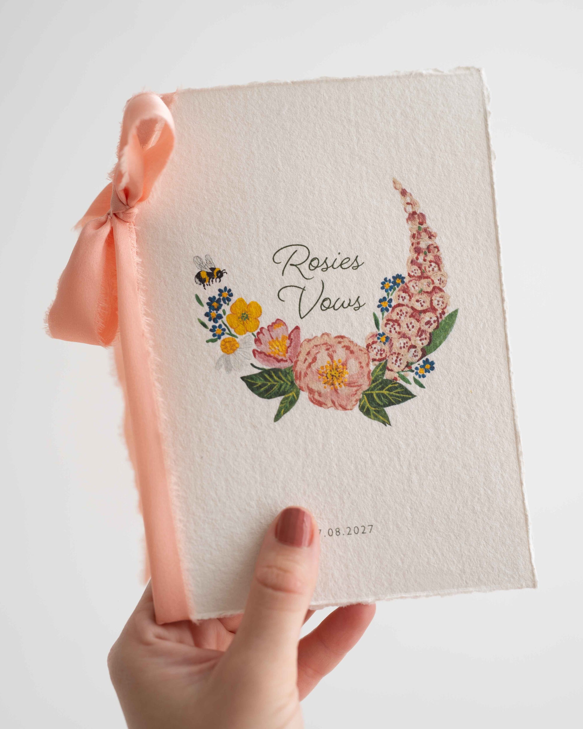 Hand holding a wild flower vow book. Watercolour hand painted florals of meadow flowers and a little bee make up a posy. Couples names are nestled in the floral with wedding date set below. Printed on handmade Indian cotton paper and styled with blush silk ribbon on a white background. Fancy Folk Design