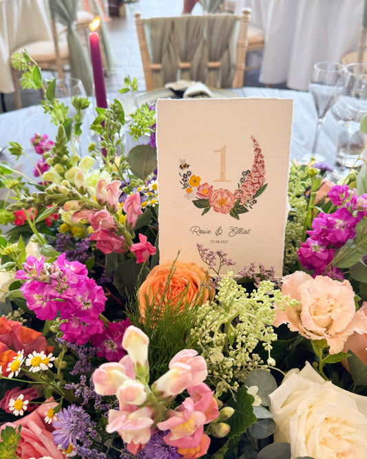 Pretty wild flower wedding table number. With gold leaf number in the centre of the posy. Little bee flying into the flowers. Couples names and wedding date printed below. Styled ona wedding breakfst table sitting inside a floral display.