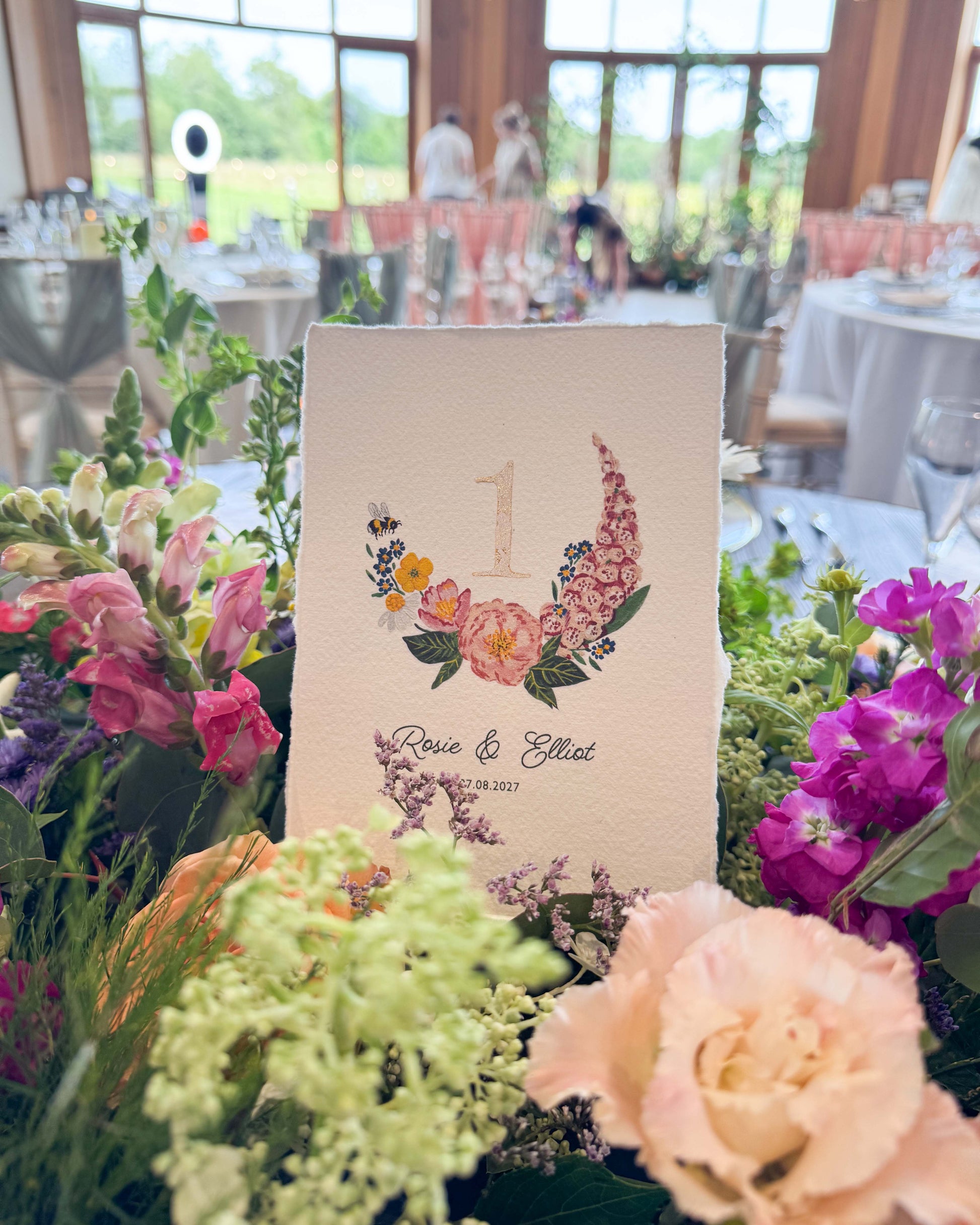Pretty wild flower wedding table number. With gold leaf number in the centre of the posy. Little bee flying into the flowers. Couples names and wedding date printed below. Styled ona wedding breakfst table sitting inside a floral display.