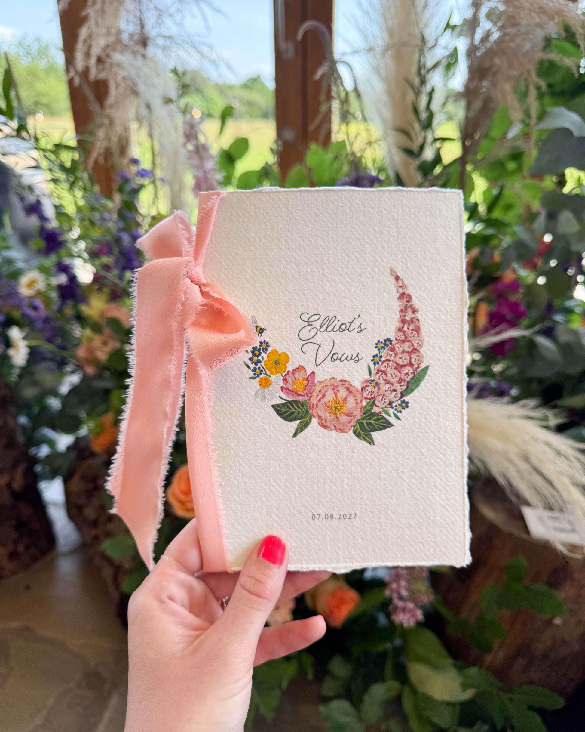 Hand holding a wild flower vow book titled 'Elliot's Vows' Wild flower Vow books. Watercolour hand painted florals of meadow flowers and a little bee make up a posy. Couples names are nestled in the floral with wedding date set below. Printed on handmade Indian cotton paper and styled with silk ribbon.  With wedding flowers in the background. Fancy Folk Design.