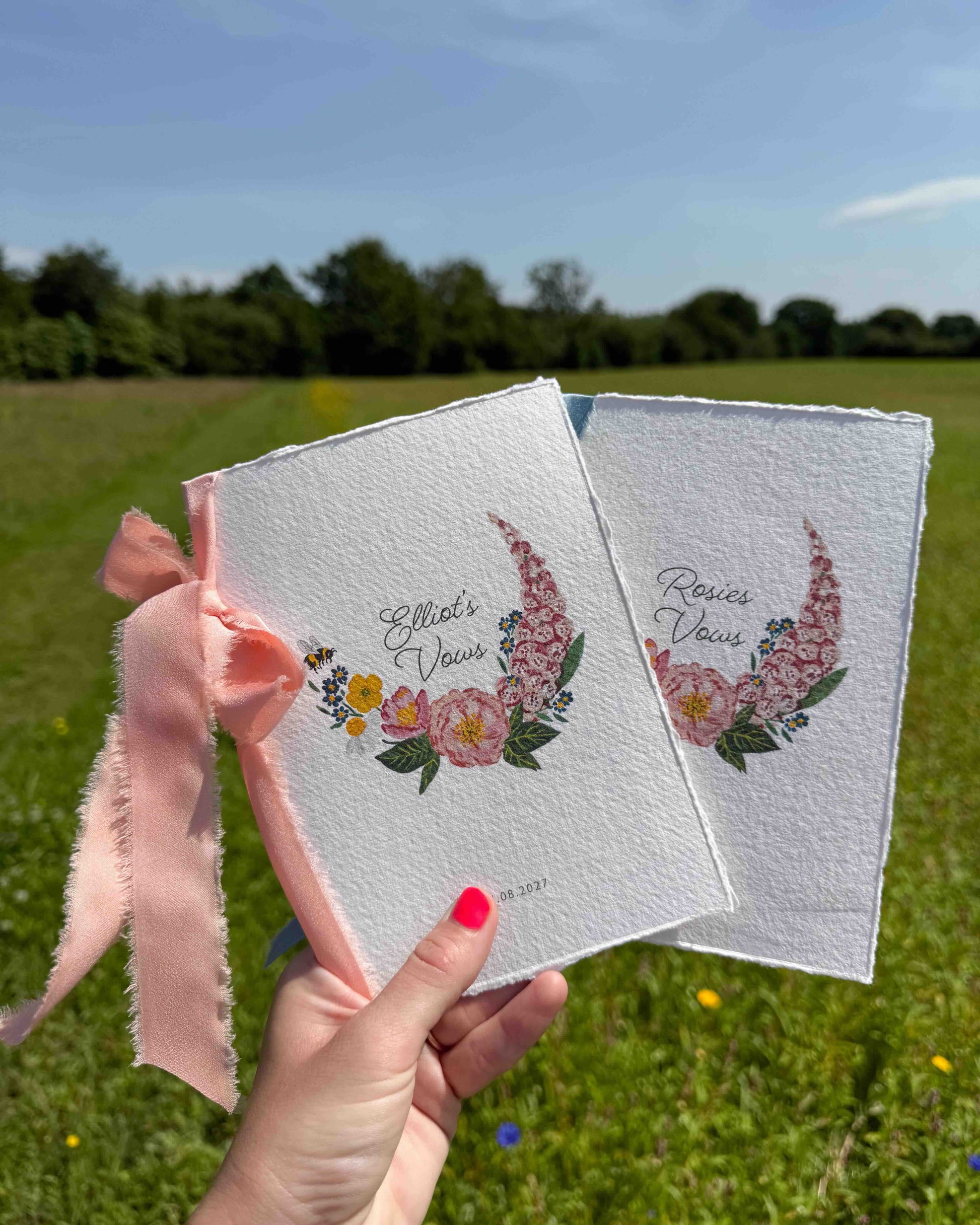 Hand holding two wild flower vow books. Watercolour hand painted florals of meadow flowers and a little bee make up a posy. Couples names are nestled in the floral with wedding date set below. Printed on handmade Indian cotton paper and styled with silk ribbon. With a wild flower meadow and mountains in the background. Fancy Folk Design