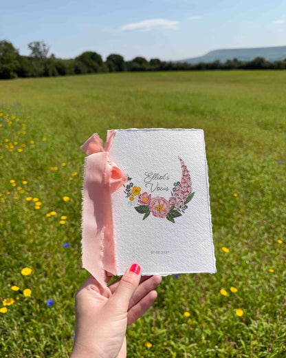 Hand holding a wild flower vow book. Watercolour hand painted florals of meadow flowers and a little bee make up a posy. Couples names are nestled in the floral with wedding date set below. Printed on handmade Indian cotton paper and styled with silk ribbon. With a wild flower meadow and mountains in the background. Fancy Folk Design