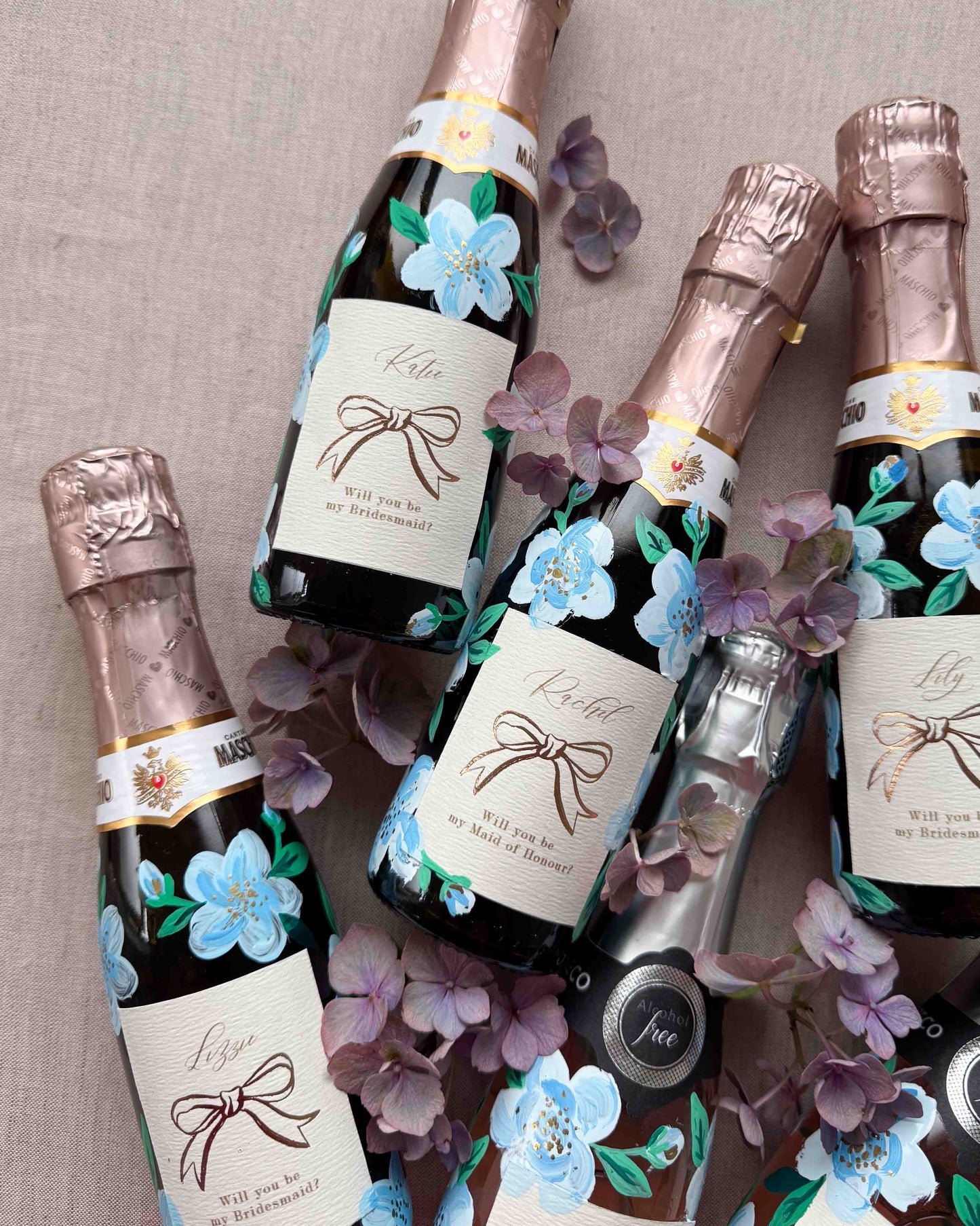 Set of mini bottles with hand painted blue florals and personalised  labels. Labels feature a foil bow design and bespoke wording - Rachel, Will you be my Maid of Honour? printed on luxurious textured card. Styled with with fresh flowers. 
