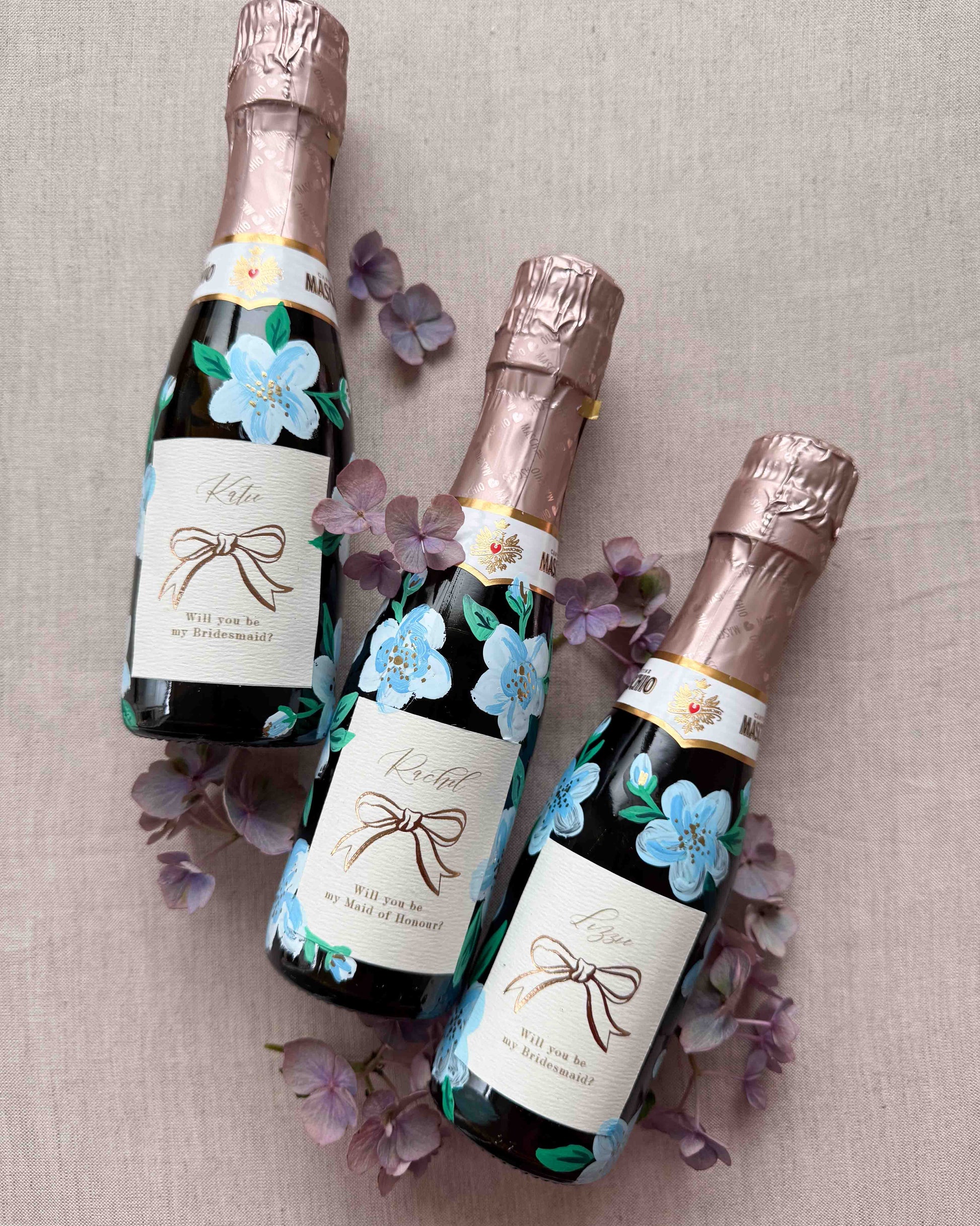 Three mini bottles with hand painted blue florals and personalised  labels. Labels feature a foil bow design and bespoke wording - Rachel, Will you be my Maid of Honour? printed on luxurious textured card. Styled with with fresh flowers. 