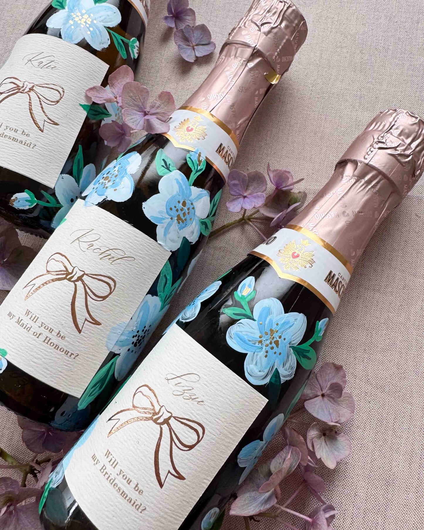 Close up of mini bottles with hand painted blue florals and personalised  labels. Labels feature a foil bow design and bespoke wording - Rachel, Will you be my Maid of Honour? printed on luxurious textured card. Styled with with fresh flowers. 