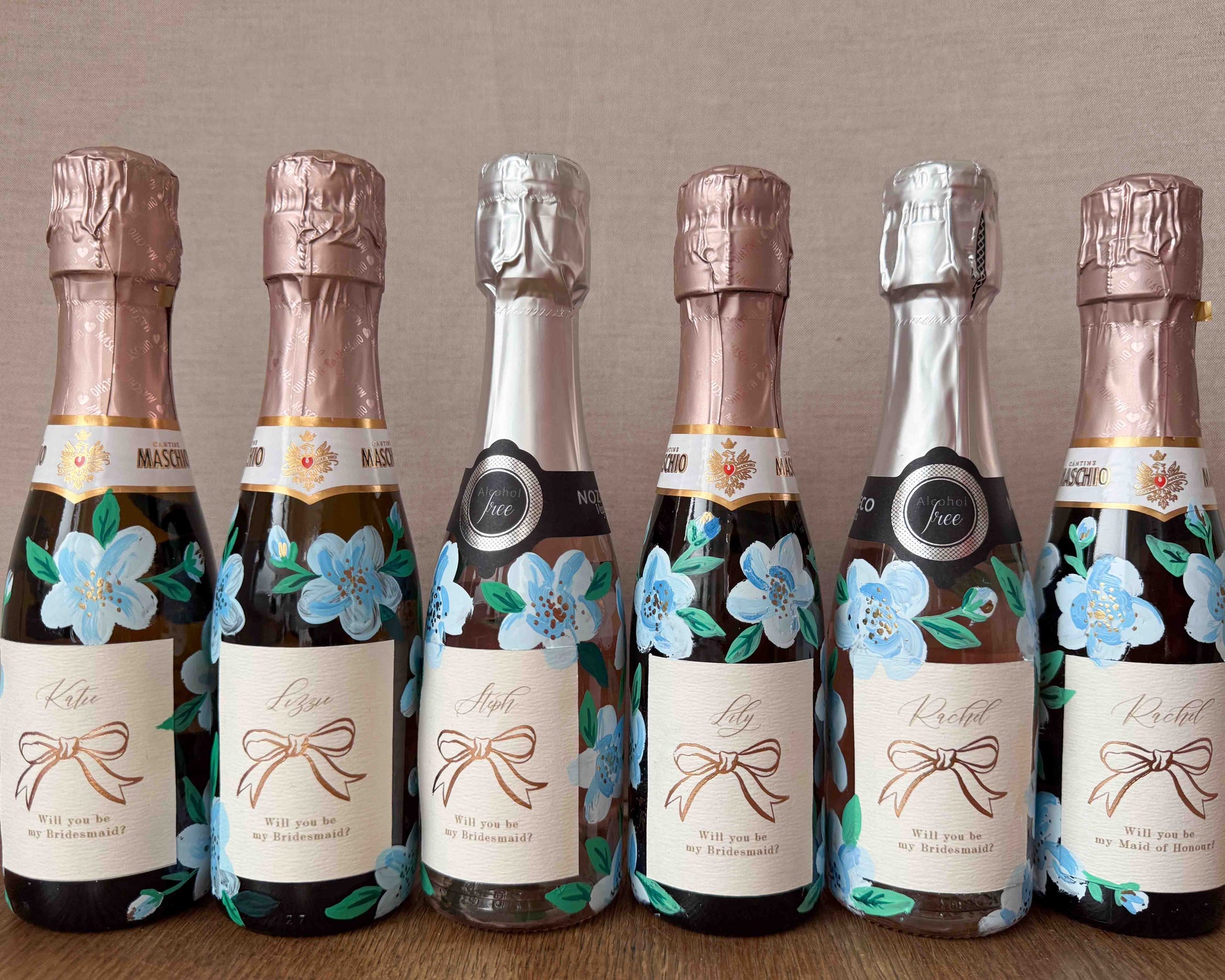 Set of mini bottles with hand painted blue florals and personalised  labels. Labels feature a foil bow design and bespoke wording - Rachel, Will you be my Maid of Honour? printed on luxurious textured card. Styled with with fresh flowers. 
