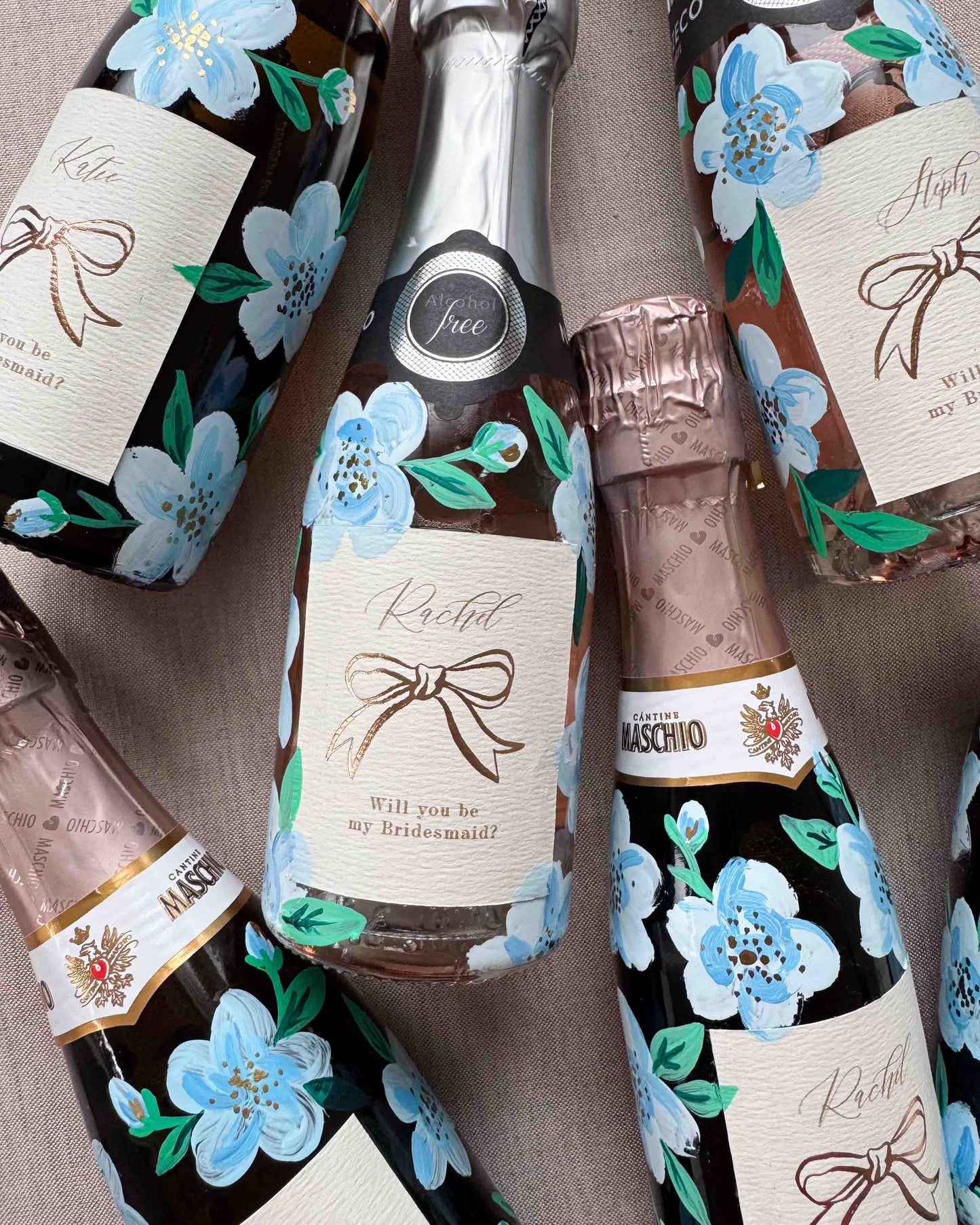 Set of mini bottles with hand painted blue florals and personalised  labels. Labels feature a foil bow design and bespoke wording - Rachel, Will you be my Maid of Honour? printed on luxurious textured card. Styled with with fresh flowers. 