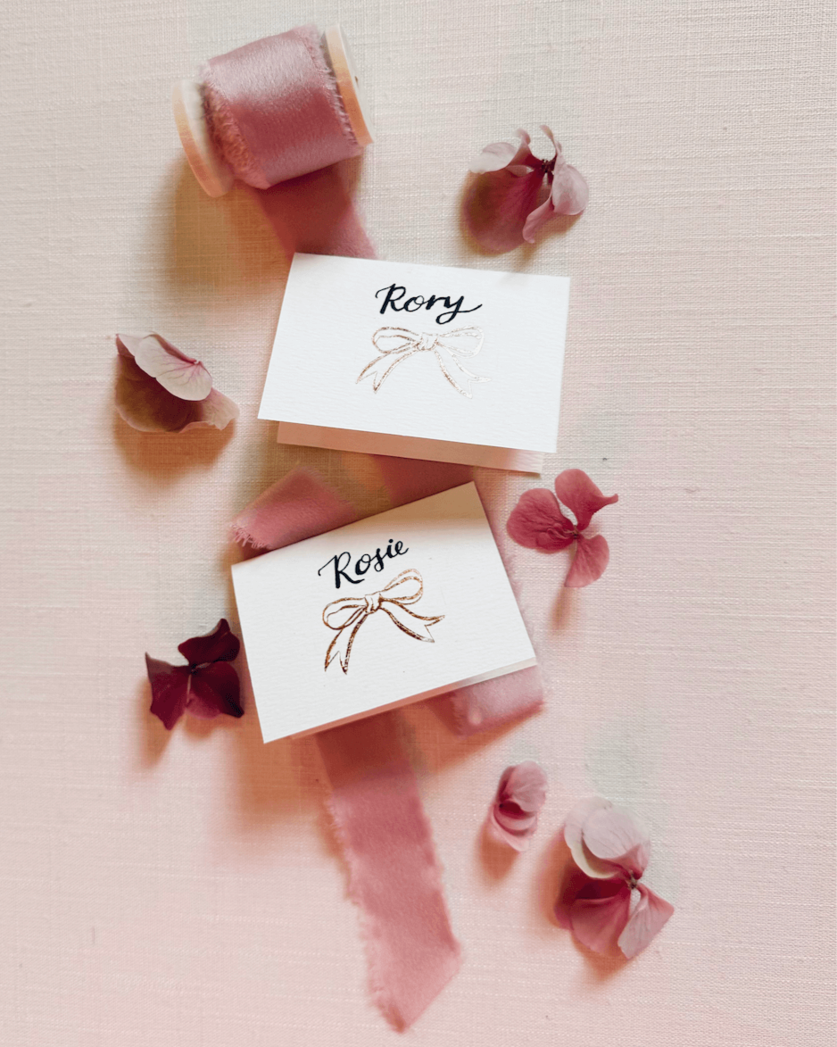 Foiled Bow design place card for yourwedding table with hand calligraphy names. Rose Gold hot foil on textured card. Styled with silk ribbon and pink flowers. DEsigned by Fancy Folk wedding stationery