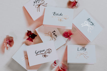 Rose gold foiled place cards for wedding table .With rose & bow icons and hand calligraphy names. Designed by Fancy Folk wedding stationery.