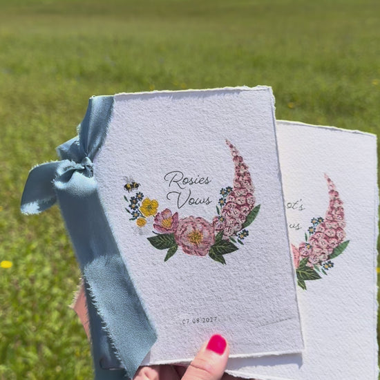 Two wild flower vow books. Watercolour hand painted florals of meadow flowers and a little bee make up a posy. Couples names are nestled in the floral with wedding date set below. Printed on handmade Indian cotton paper and styled with silk ribbon. With a wild flower meadow and mountains in the background. Fancy Folk Design