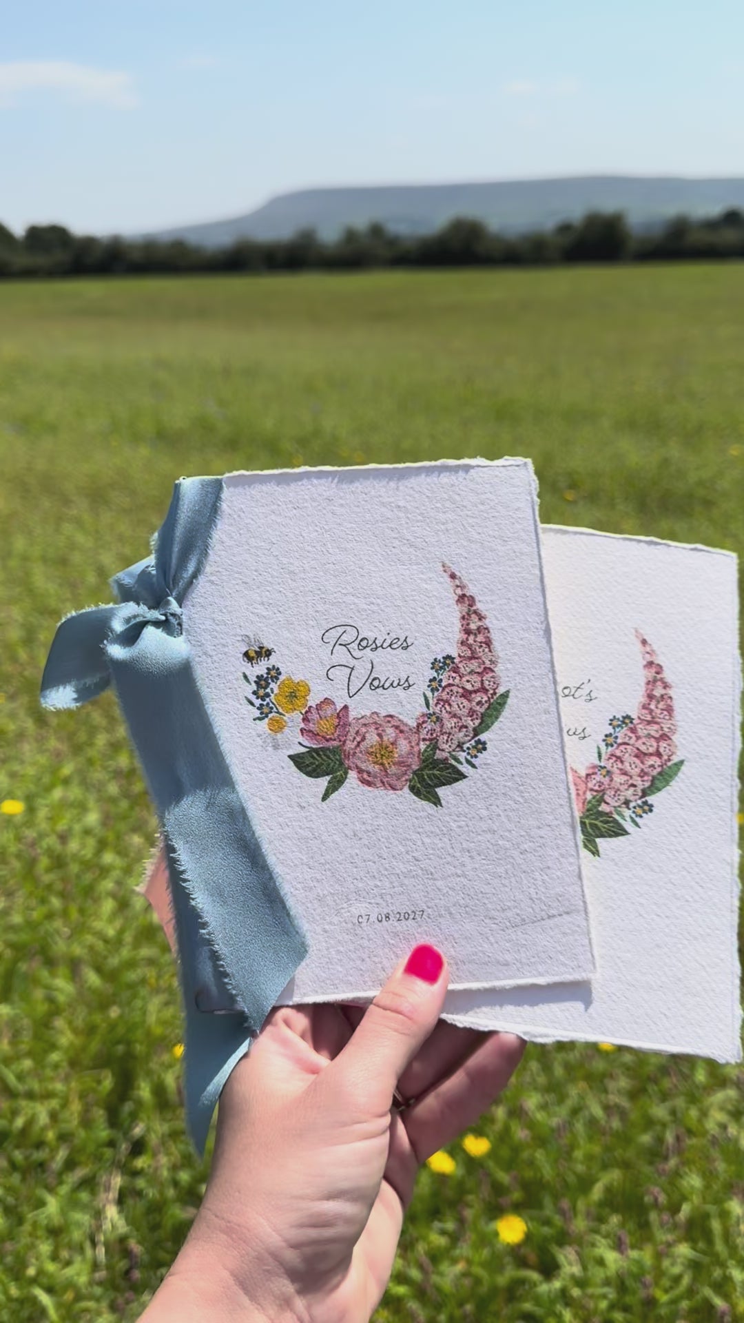 Two wild flower vow books. Watercolour hand painted florals of meadow flowers and a little bee make up a posy. Couples names are nestled in the floral with wedding date set below. Printed on handmade Indian cotton paper and styled with silk ribbon. With a wild flower meadow and mountains in the background. Fancy Folk Design
