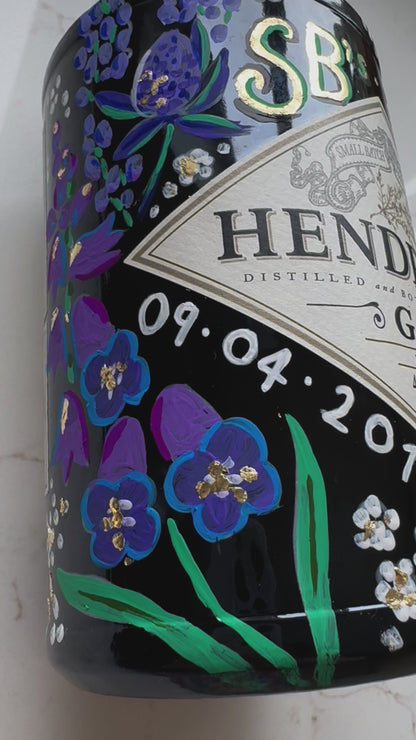 close up video of hand painted occasion bottle. Blue and Purple Wildflowers which are embellished with gold leaf. Personalised initials and dates.