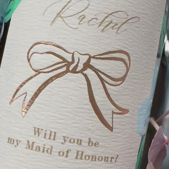 Hand painted blue flowers with gold accents in the centres. Painted onto mini bottles of Prosecco. With personalised label, writing ready Rachel, will you be my Maid of Honour? with a rose gold bow in the centre. A perfect bridesmaids proposal gift. Fancy Folk Design