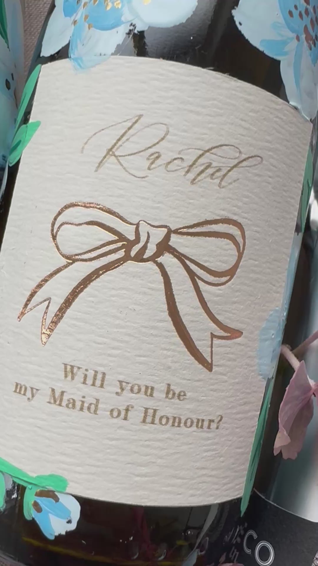 Hand painted blue flowers with gold accents in the centres. Painted onto mini bottles of Prosecco. With personalised label, writing ready Rachel, will you be my Maid of Honour? with a rose gold bow in the centre. A perfect bridesmaids proposal gift. Fancy Folk Design