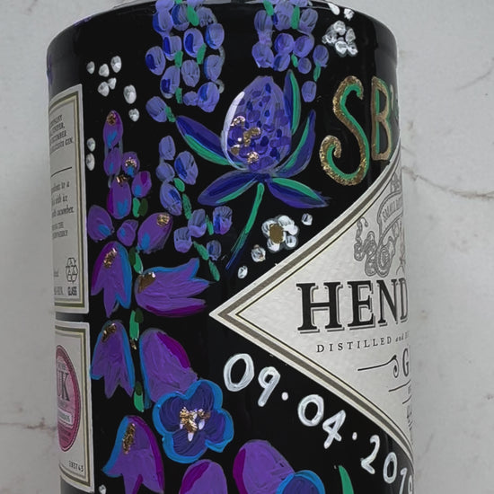 Close up video of hand painted occasion bottle. Blue and Purple Wildflowers which are embellished with gold leaf. Personalised initials and dates.