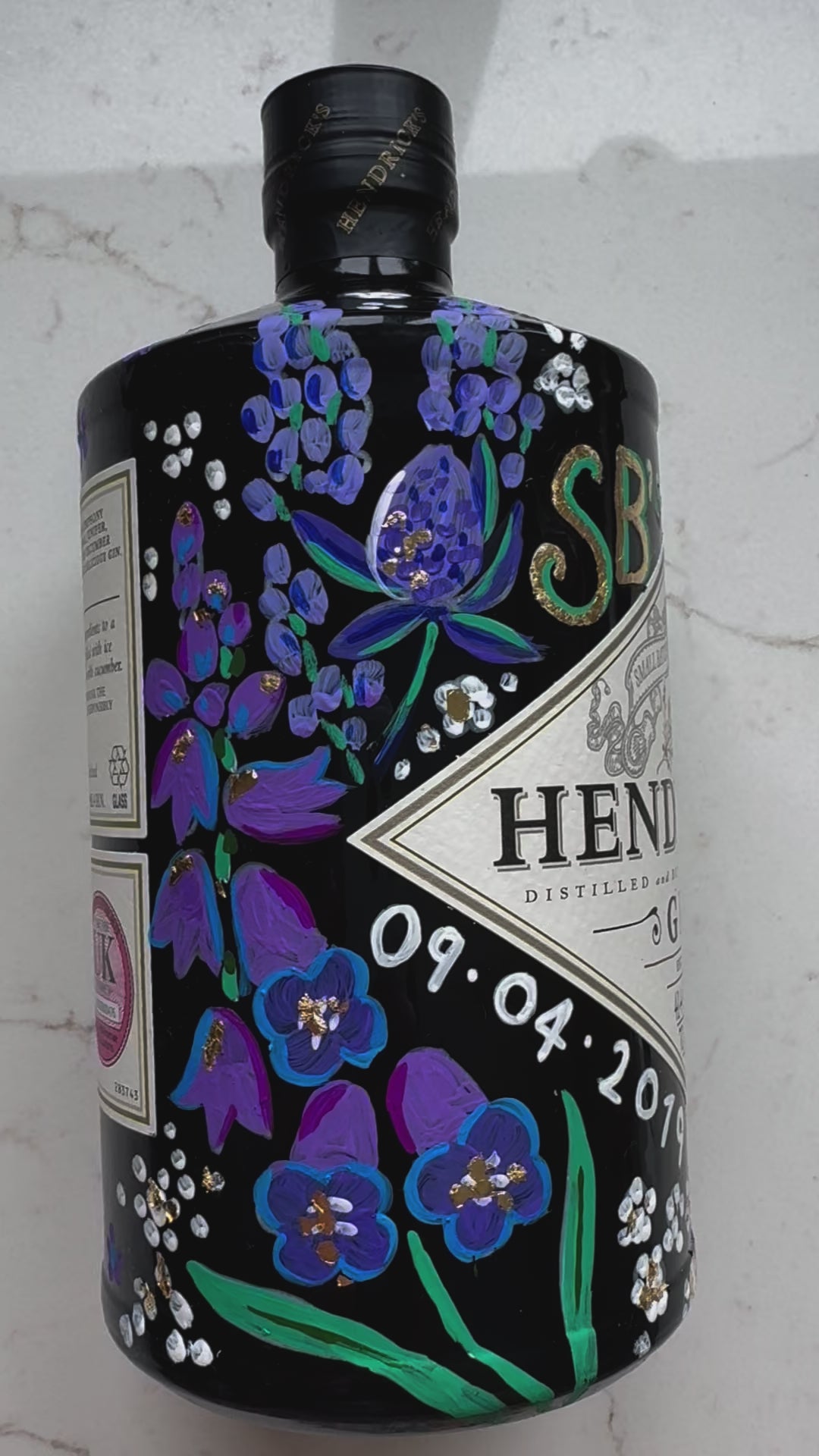 Close up video of hand painted occasion bottle. Blue and Purple Wildflowers which are embellished with gold leaf. Personalised initials and dates.