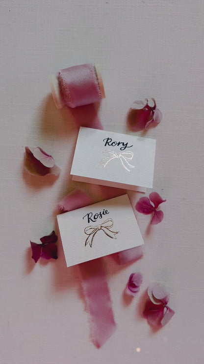 Foiled Bow design place card for your wedding table with hand calligraphy names. Styled with silk ribbon & pink flowers. Designed by Fancy Folk wedding stationery