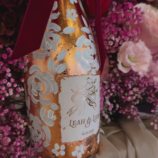  Hand painted wedding Champagne Bottle. Lux Copper leaf base with white painted roses all around the bottle. Personalise label with couples names and wedding date. Perfect for a Champagne Tower moment. Pink florals in the background. By Fancy Folk Design