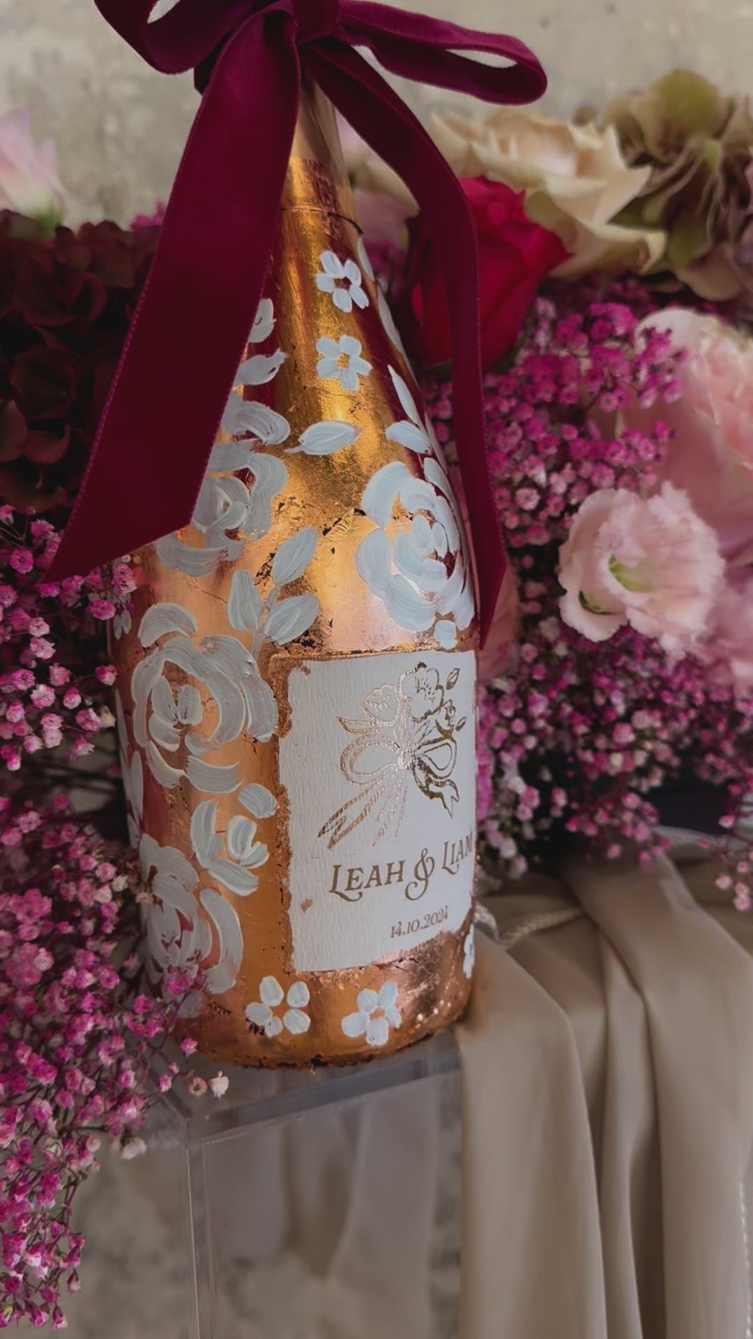  Hand painted wedding Champagne Bottle. Lux Copper leaf base with white painted roses all around the bottle. Personalise label with couples names and wedding date. Perfect for a Champagne Tower moment. Pink florals in the background. By Fancy Folk Design