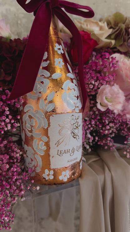  Hand painted wedding Champagne Bottle. Lux Copper leaf base with white painted roses all around the bottle. Personalise label with couples names and wedding date. Perfect for a Champagne Tower moment. Pink florals in the background. By Fancy Folk Design
